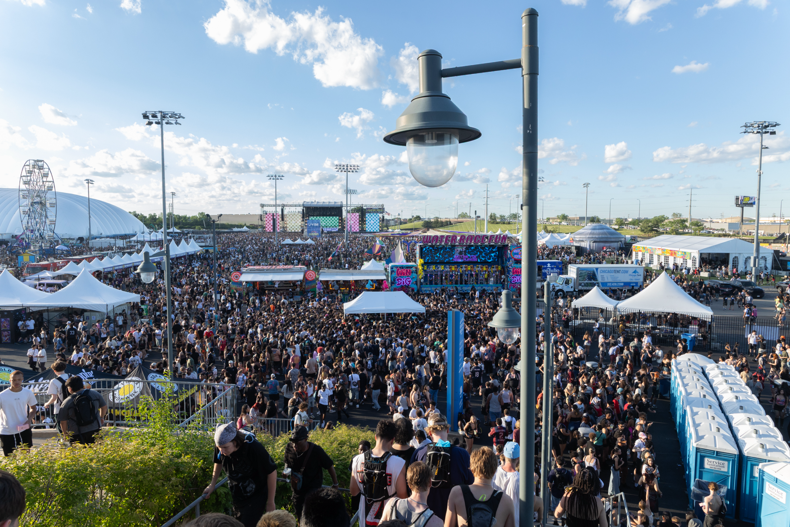 Elevated view of thousands of attendees at the Summer Smash music festival, with the main stage, food vendors, and white event tents spread across the festival grounds.