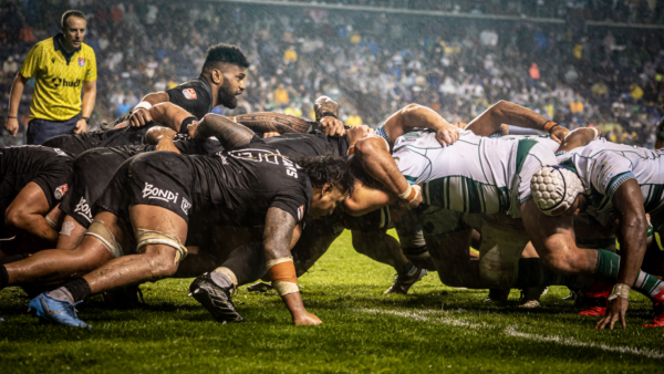 Rugby players from opposing teams compete in a scrum during a rainy night match in front of a packed crowd.