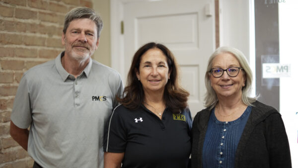Three adults stand side by side, smiling at the camera in an office with exposed brick walls and a doorway in the background.