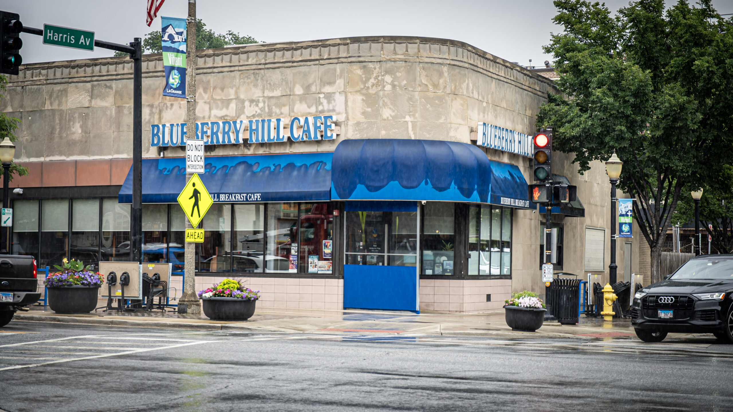 A corner view of Blueberry Hill Cafe on a rainy day, with blue awnings, wet streets, a red traffic light, crosswalk signs, and flower planters at the intersection.