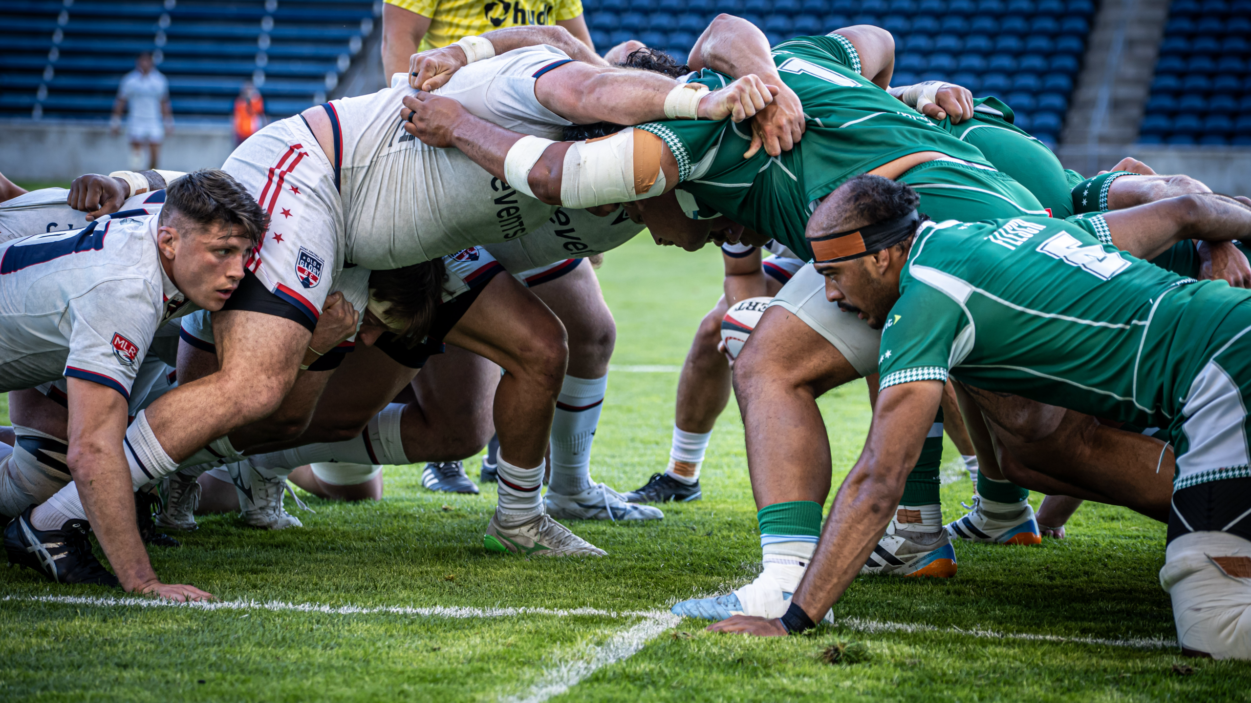 Two rugby teams engage in a scrum during a match, with players in white and green jerseys pushing against each other on the field.