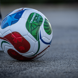 Multi-color Fifa 2026 soccer ball seen on pavement with pair of blue and white shoes behind it