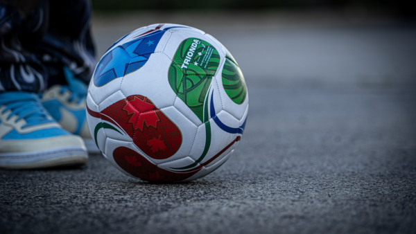 Multi-color Fifa 2026 soccer ball seen on pavement with pair of blue and white shoes behind it