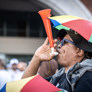 A fan wearing a rainbow-colored umbrella hat blows an orange horn while cheering before the game in the parking lot.