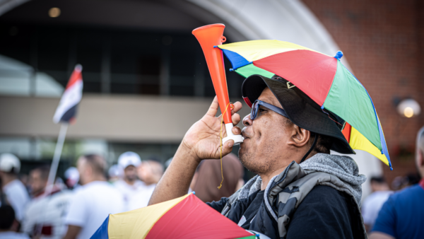 A fan wearing a rainbow-colored umbrella hat blows an orange horn while cheering before the game in the parking lot.