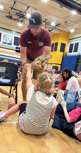 Presenter holds an iguana as children sit on a gym floor and reach out to touch it during an educational program.