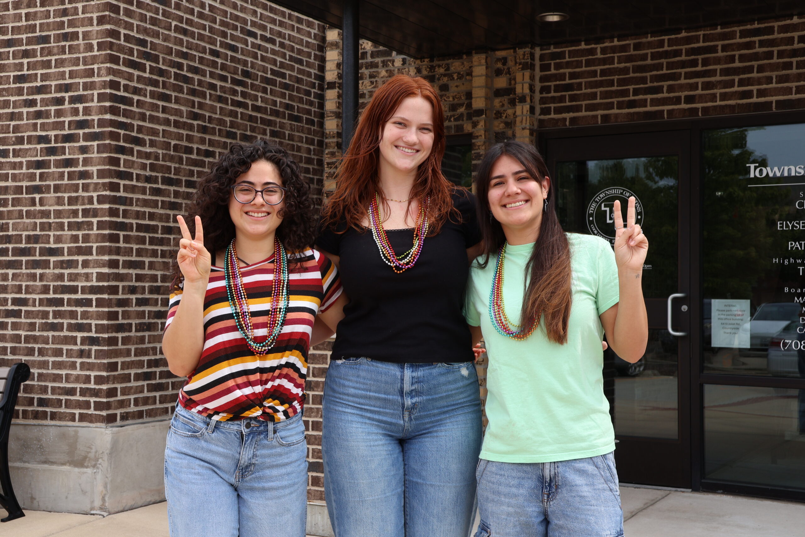 Three smiling people stand arm in arm outside a brick building, wearing colorful bead necklaces. The two people on either side are making peace signs toward the camera.