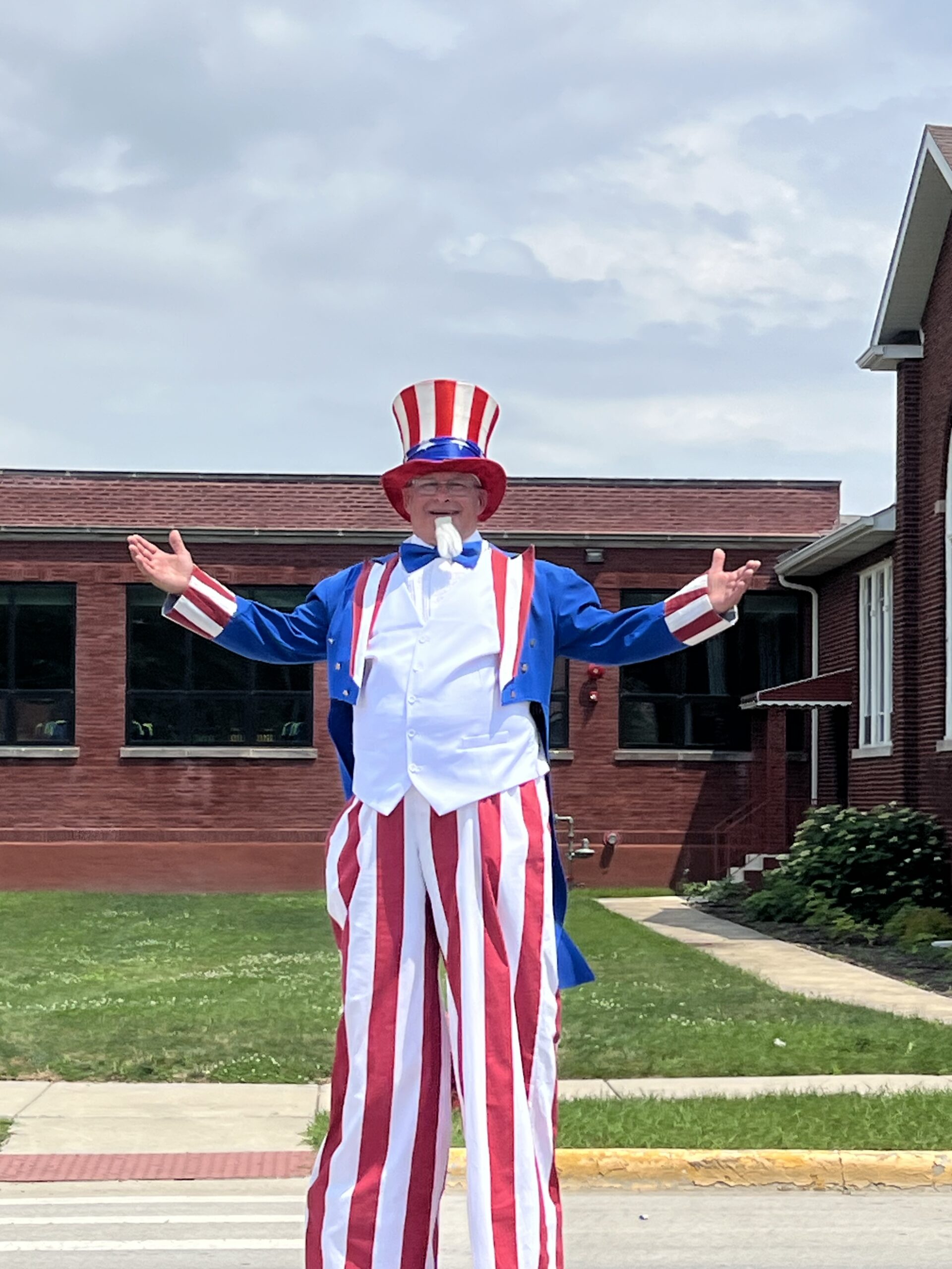 A smiling person dressed as Uncle Sam stands on stilts with their arms outstretched during a Fourth of July parade. He is wearing a tall red, white, and blue striped hat, a blue jacket, a white vest, red-and-white striped pants that cover the stilts. He is in front of a brick building and it is cloudy outside.