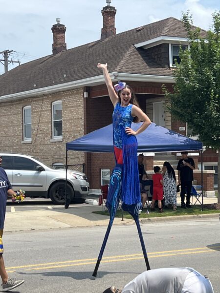 Stilt walker in a blue costume waves while performing in a Fourth of July parade.