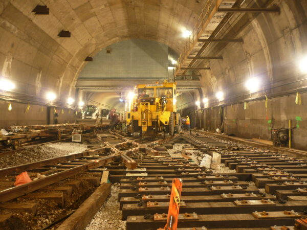 Construction crews work inside a large transit tunnel using heavy equipment under bright work lights.