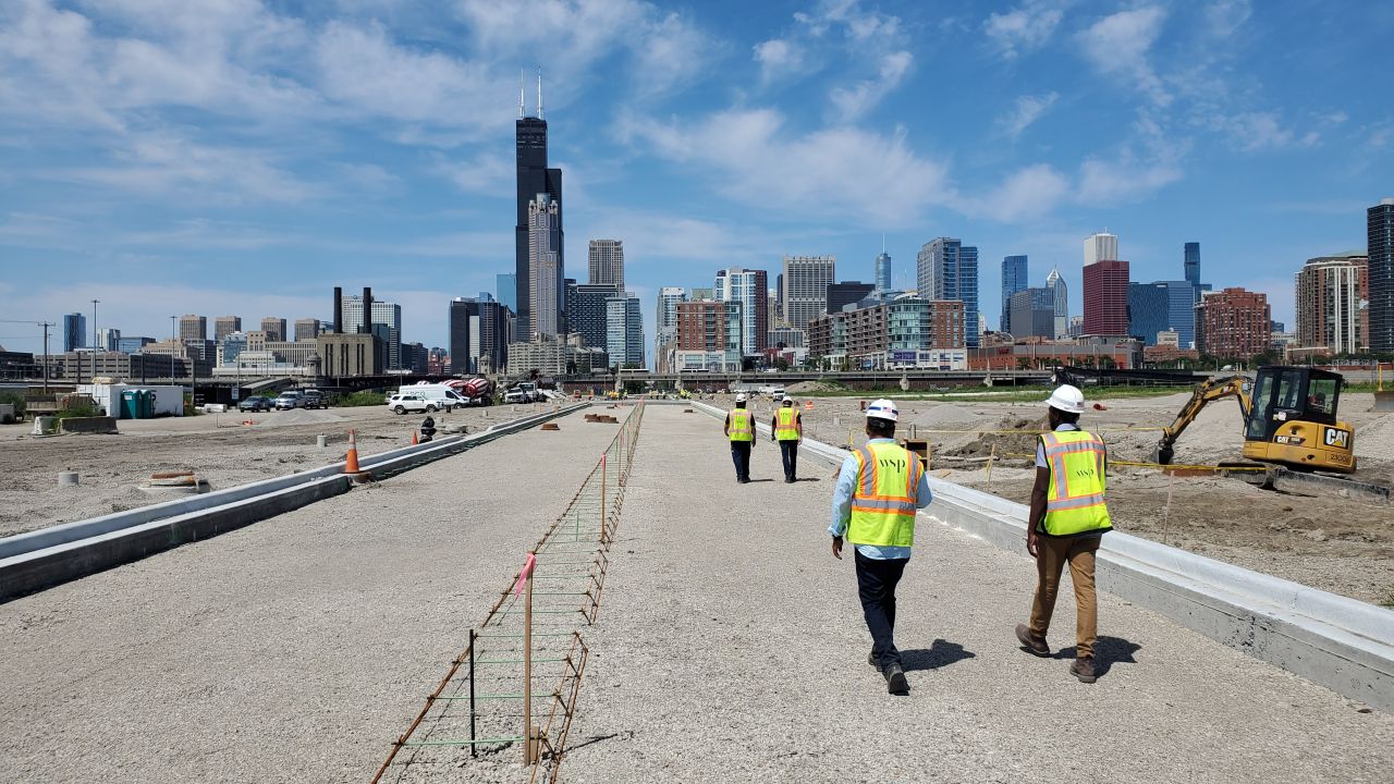 Construction workers walk along a newly built roadway toward the Chicago skyline under a bright blue sky.