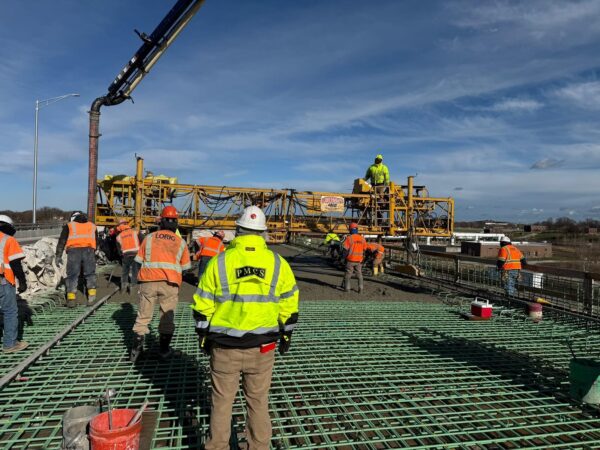 Construction workers pour concrete on a bridge deck over steel reinforcement using a concrete pump on a clear day.