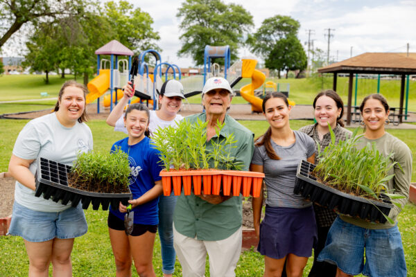 Seven volunteers pose together at a community garden, smiling while holding trays of seedlings and native plants. A playground and picnic shelter are visible in the background.