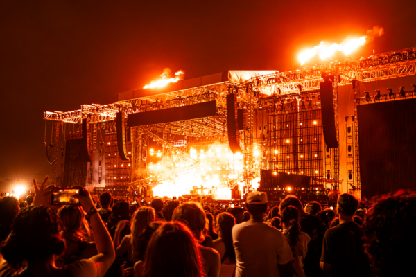Crowd watching a Skrillex's set with bright orange stage lights and pyrotechnic flames above the stage.