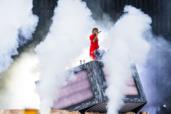 Baby Keem wearing a bright red outfit sings into a microphone while standing on an elevated stage platform surrounded by large clouds of white smoke.