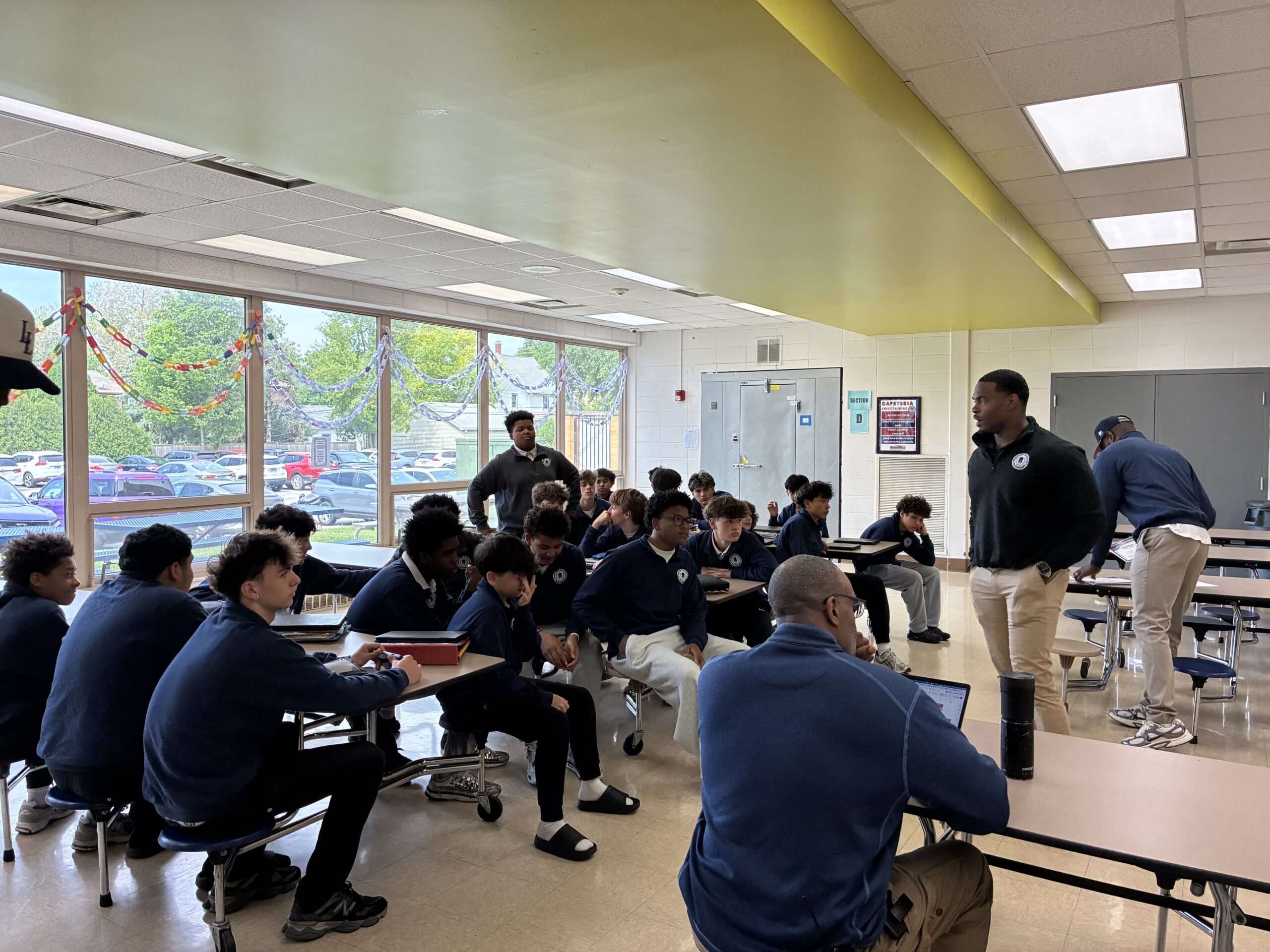 Students in a school cafeteria listen as staff members lead a group discussion during a session of the men's mental health warrior academy during school.