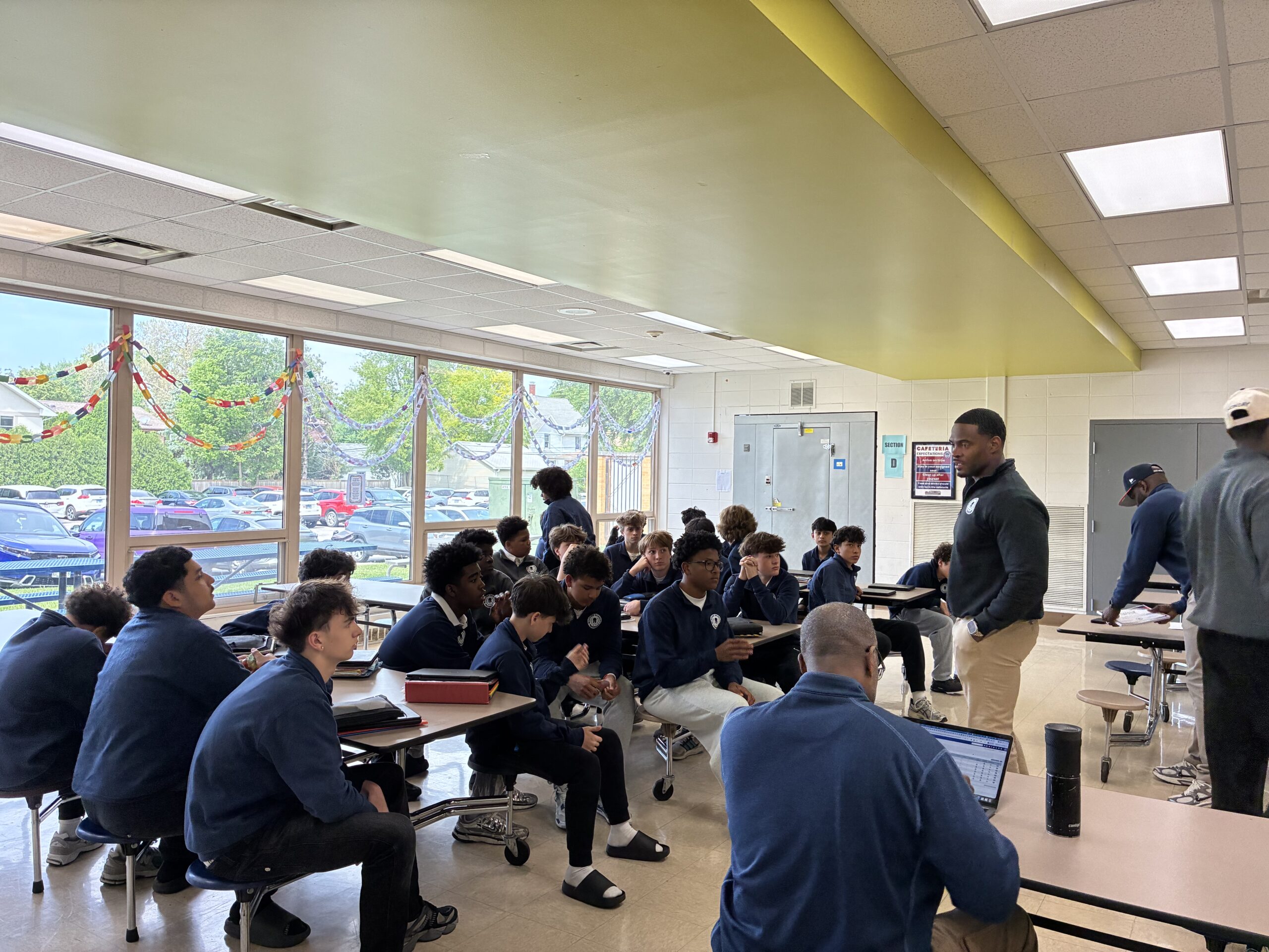 Students in a school cafeteria listen as staff members lead a group discussion during a session of the men's mental health warrior academy during school.