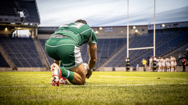 Rugby player kneels to set up a kick during a match, with teammates and goalposts visible in the background.
