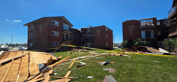 Three brick apartment buildings show extensive storm damage, including a collapsed roof on two of the buildings, destroyed upper-floor balconies, and exposed interiors. Roofing materials and wood debris cover the grass lawn while yellow caution tape blocks access to the damaged area.