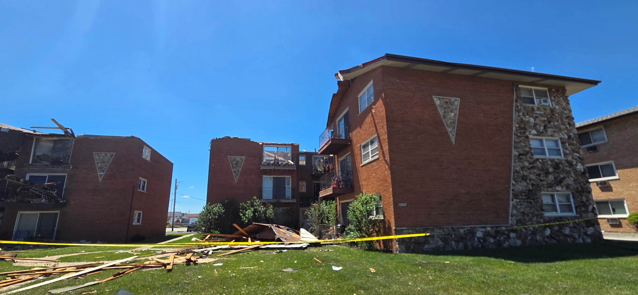 Three brick apartment buildings with severe storm damage, including missing roof sections and destroyed balconies on the upper floors. Debris is scattered across the lawn and yellow caution tape blocks off access to the damaged area.