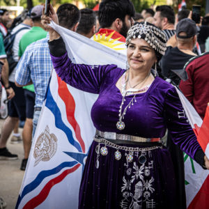 A person in a purple traditional outfit and silver headpiece smiles while holding a flag at a soccer game, surrounded by fans carrying Iraqi flags.