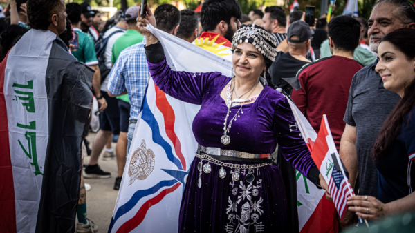 A person in a purple traditional outfit and silver headpiece smiles while holding a flag at a soccer game, surrounded by fans carrying Iraqi flags.