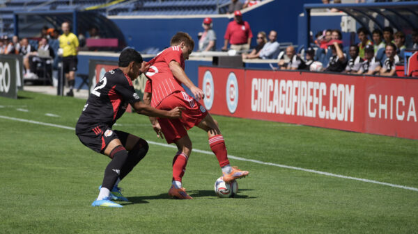 Chicago Fire II player in a red uniform shields the ball from a Red Bull New York II player in a black uniform near the sideline.