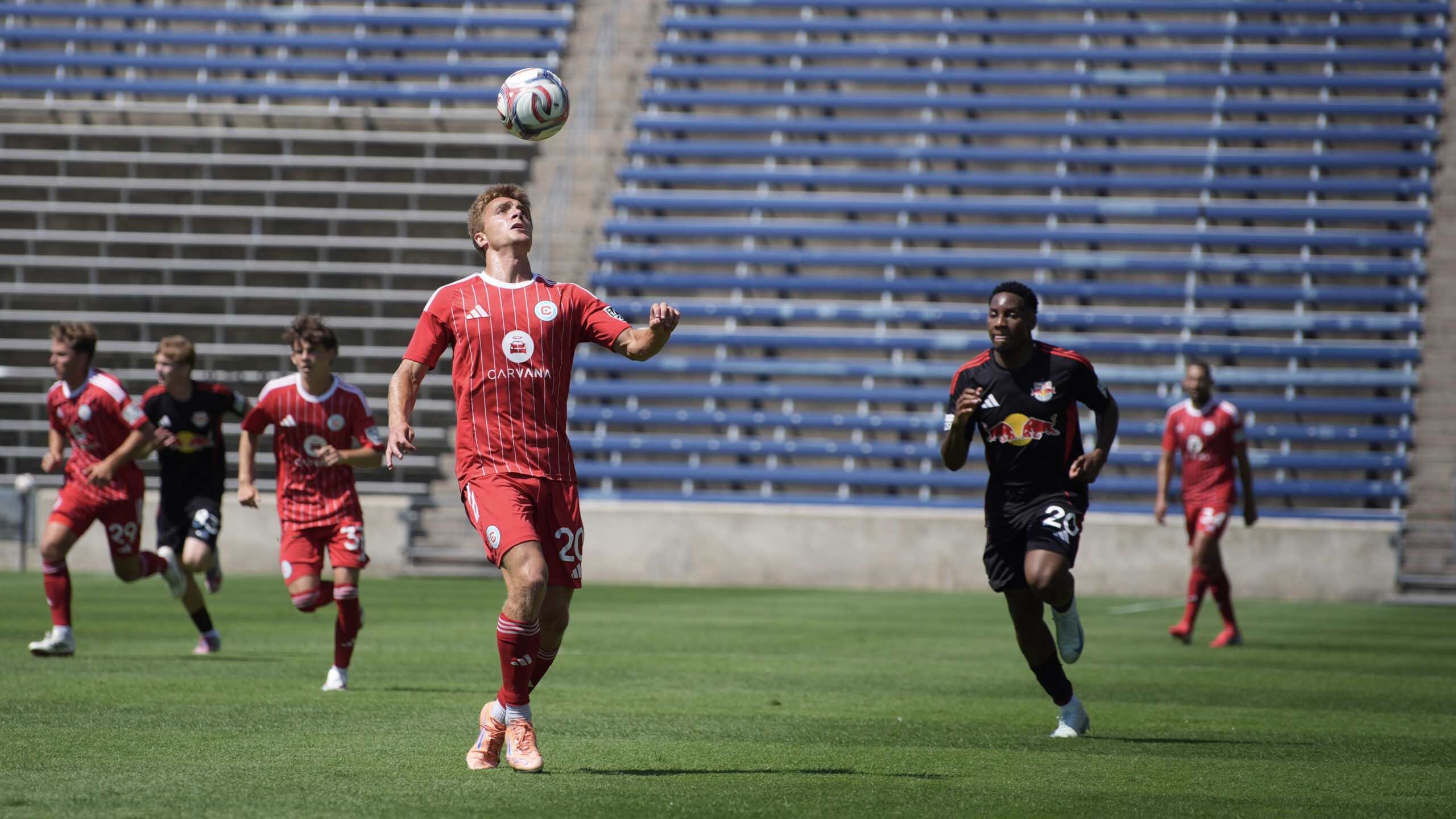 Chicago Fire II player in a red uniform runs toward a ball in the air preparing to take a header, with several opposing players chasing behind him on a field.