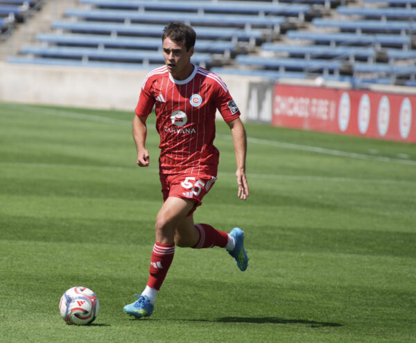 Chicago Fire II player in a red uniform runs with the ball across the field, with empty blue bleachers and a sideline advertisement in the background.