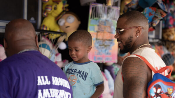 A young boy wearing a Spider-Man shirt stands between two adults at a carnival booth. One man wears a purple shirt, while the other wears a white shirt, glasses, a gold chain and a Spider-Man backpack. Colorful stuffed animals and toys fill the background.