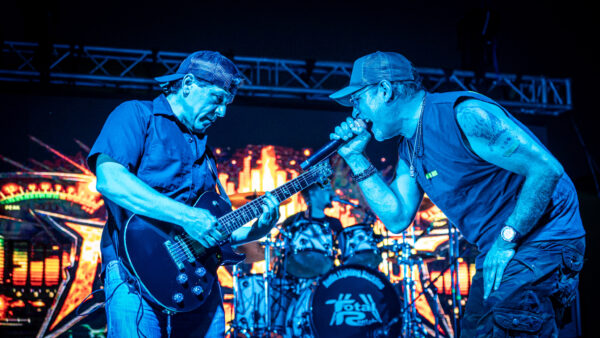 Two musicians perform onstage under vivid blue lighting. One plays an electric guitar while the other sings into a microphone. A drum set and colorful stage lights are visible in the background.