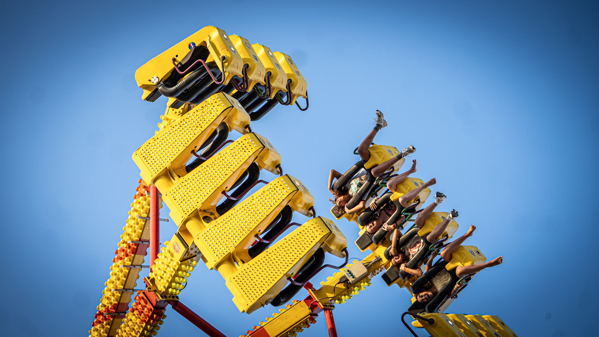 A group of people ride a yellow amusement park ride that swings high into the air against a clear blue sky. Riders are secured in seats along the ride’s arms, with several raising their legs as the ride tilts upward.
