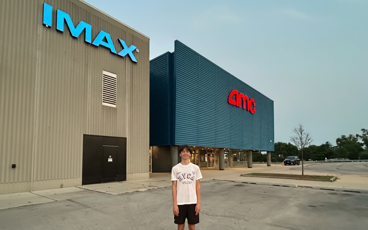 Young man stands in front of an IMAX AMC movie theater on a day with clear skys.