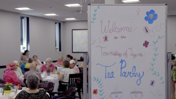 A white board with drawings and the words “Welcome to the Township of Lyons Tea Party.” Tea party attendees are seen behind the sign sitting at tables and talking to one another.