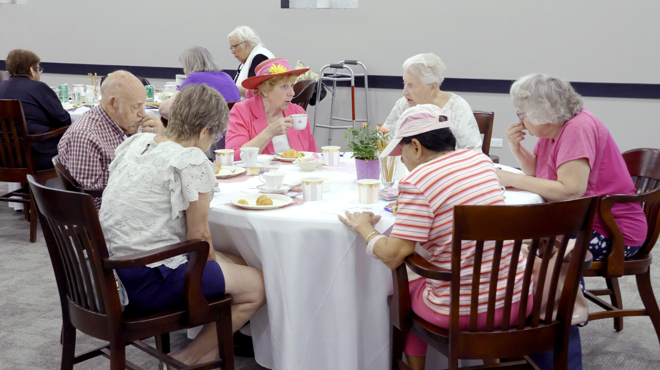 A group of older adults gathers around a table for tea and pastries. They sit in a bright community room, with cups, plates and a small floral centerpiece on the table. One person wears a colorful flowered hat, and a walker is visible in the background.
