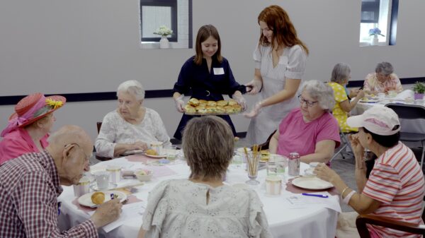 Two young women serve sandwiches to a group of older adults gathered around a table for tea. The table is set with plates, cups, pastries and drinks, while other guests sit at tables in the background. One woman wears a bright flowered hat.
