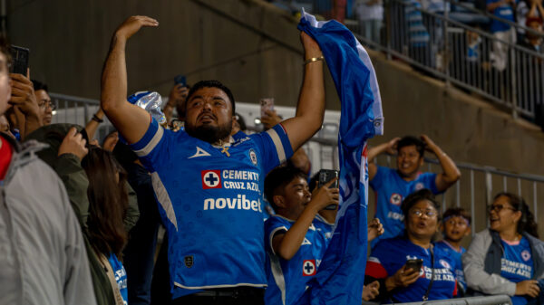 A Cruz Azul fan in a blue jersey raises his arms and holds a blue-and-white shirt overhead while surrounded by cheering supporters.