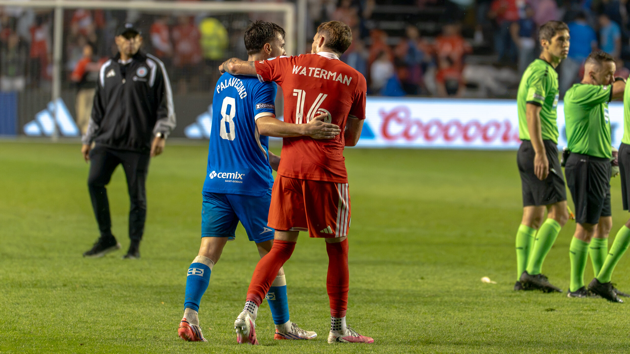 Two soccer players in blue and red jerseys walk together with their arms around each other on the field, while referees and spectators appear in the background.