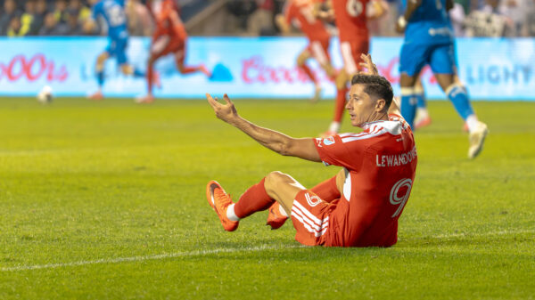 A soccer player in a red uniform sits on the field with his arms raised, appearing to protest a call during the match.