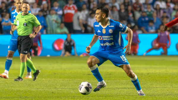 A Cruz Azul player in a blue uniform runs with the ball during a soccer match, with a referee, other players, and spectators in the background.