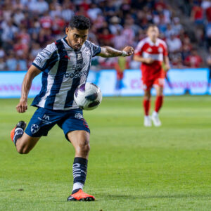 A soccer player in a black-and-white uniform runs while controlling the ball during a match, with other players and spectators in the background.