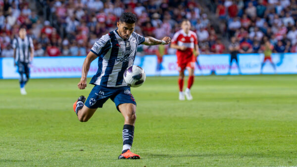 A soccer player in a black-and-white uniform runs while controlling the ball during a match, with other players and spectators in the background.