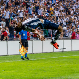 A soccer player in a black-and-white uniform performs a dramatic midair stunt in front of a large cheering crowd, with an assistant referee nearby.