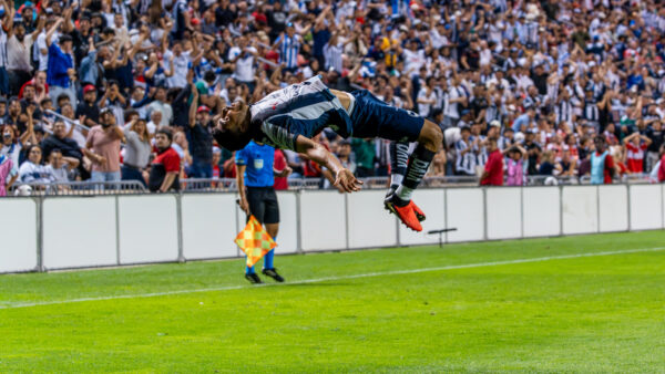 A soccer player in a black-and-white uniform performs a dramatic midair stunt in front of a large cheering crowd, with an assistant referee nearby.