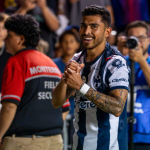 A smiling Monterrey player claps near the corner flag while spectators and security staff watch from behind.