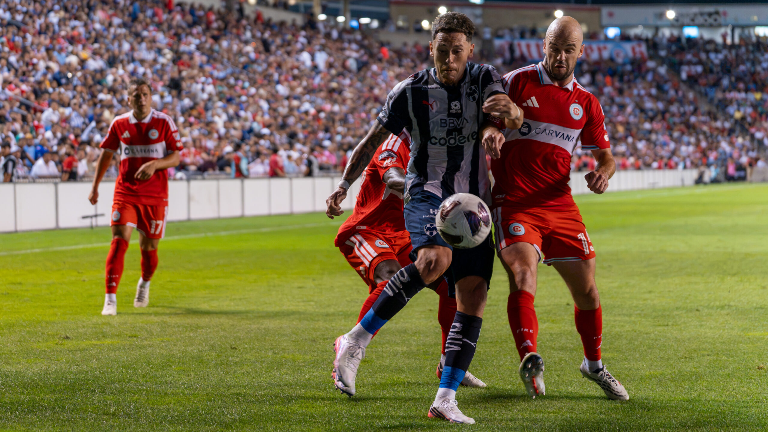 Soccer players in red and black-and-white uniforms battle for the ball on the field in front of a packed stadium.